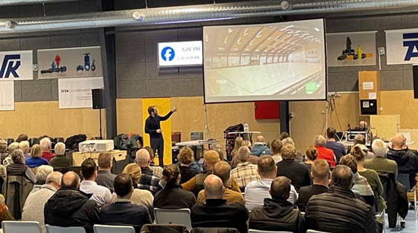 En mand peger på et slideshow i en idrætshal foran en stor forsamling siddende på stole i hallen.